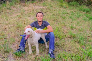 Parktaki çimlerin üzerinde oturan laggoto-Romagnolo cinsinden beyaz bir köpeği olan bir adam. Parkta köpeği gezdiriyorum. Mantar arayan köpek.