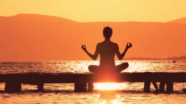Doğadaki Yoga. Bu video siluetli genç bir bayanın ahşap iskelede yoga yaptığını gösteriyor. Güneş batıyor ve çevre turuncu parlak bir ışıkla yıkanmış. Bu klip rahatlatıcı bir ruh hali ve ruhani olmak için mükemmel.. 