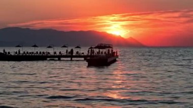 Tanınamaz haldeki insan siluetleri turuncu gökyüzüne karşı iskelede yürüyor ve güneş batarken Karadeniz üzerinde batıyor. Yaz, tropik, deniz manzarası, tatil ve doğa konsepti