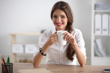 Genç bir iş kadını ofisinde fincanla masada oturuyor.