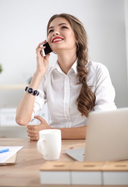 Genç iş kadını masada oturuyor ve telefonla konuşuyor.