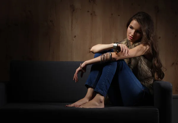 Porträt einer eleganten Frau, die auf einem schwarzen Sofa sitzt und eine blaue Jeans und Pelzweste trägt — Stockfoto