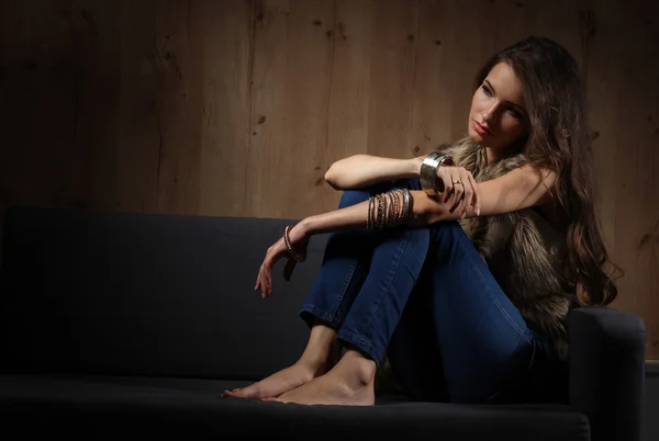 Portrait of elegant woman sitting on black sofa wearing a blue jeans and fur vest — Stock Photo, Image