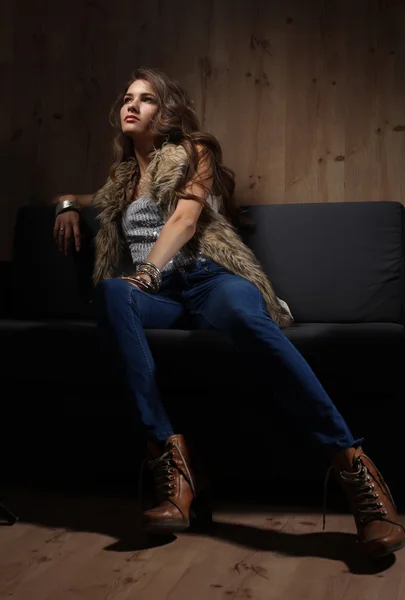 Retrato de una mujer elegante sentada en un sofá negro con pantalones vaqueros azules y chaleco de piel — Foto de Stock