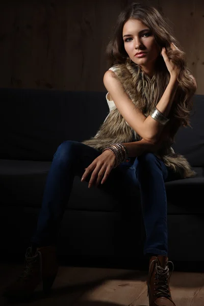 Portrait of elegant woman sitting on black sofa wearing a blue jeans and fur vest — Stock Photo, Image