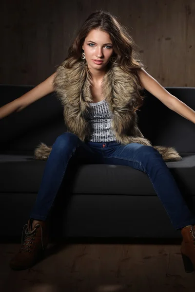 Portrait of elegant woman sitting on black sofa wearing a blue jeans and fur vest — Stock Photo, Image
