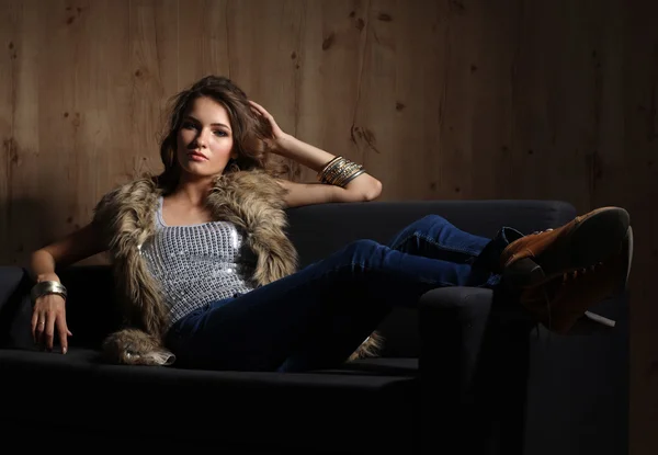 Porträt einer eleganten Frau, die auf einem schwarzen Sofa sitzt und eine blaue Jeans und Pelzweste trägt — Stockfoto