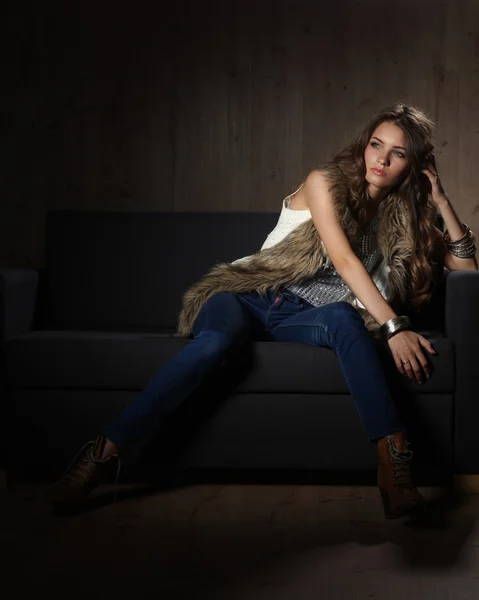 Retrato de mulher elegante sentado no sofá preto vestindo uma calça azul e colete de pele — Fotografia de Stock