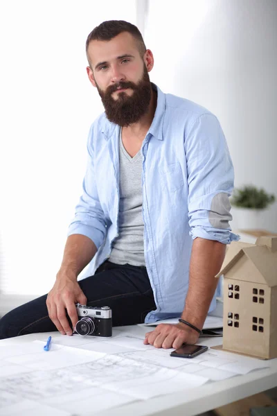 Portret van mannelijke ontwerper zittend op een tafel met blauwdrukken en vasthouden van camera — Stockfoto