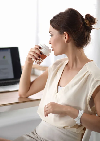 Приваблива жінка сидить за столом в офісі, працює з ноутбуком, виймаючи каву — стокове фото