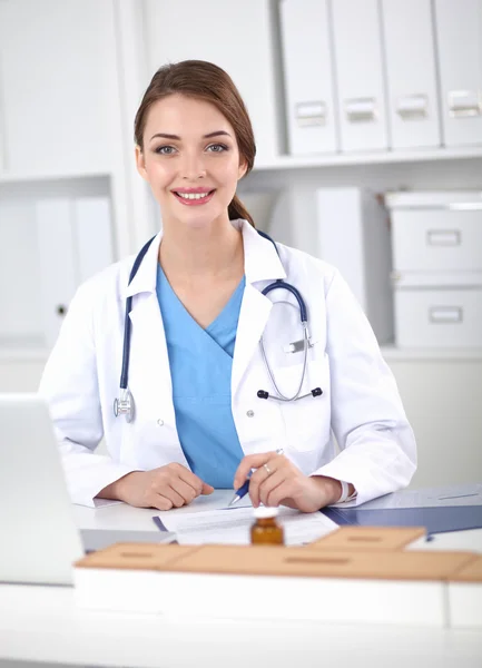 Hermosa joven sonriente doctora sentada en el escritorio y escribiendo. —  Fotos de Stock