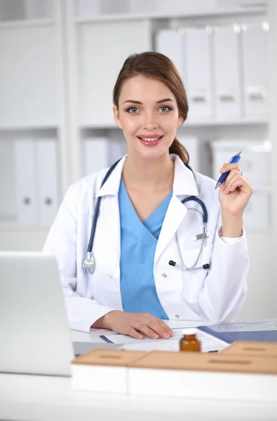 Mooie jonge glimlachende vrouwelijke arts zitten aan het bureau en schrijven. — Stockfoto