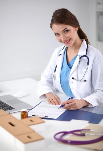 Hermosa joven sonriente doctora sentada en el escritorio y escribiendo. — Foto de Stock