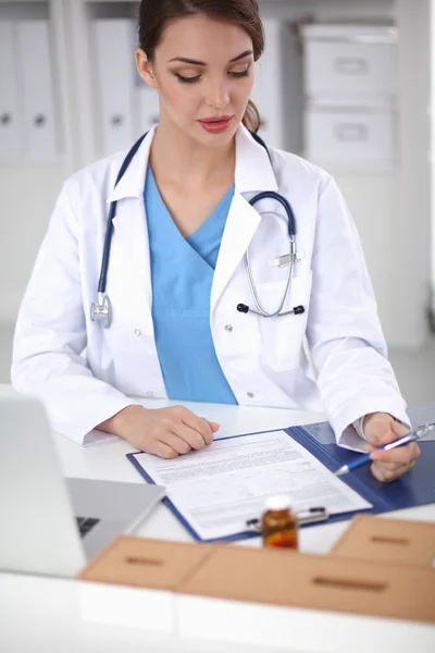 Schöne junge lächelnde Ärztin sitzt am Schreibtisch und schreibt. — Stockfoto