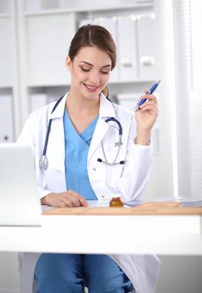 Hermosa joven sonriente doctora sentada en el escritorio y escribiendo. — Foto de Stock