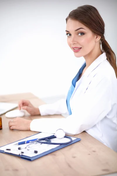 Hermosa joven sonriente doctora sentada en el escritorio y escribiendo. — Foto de Stock