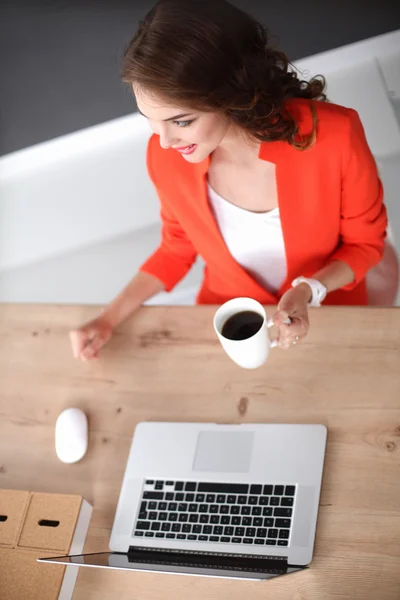 Mujer atractiva sentada en el escritorio en la oficina, trabajando con el ordenador portátil, sosteniendo una taza —  Fotos de Stock