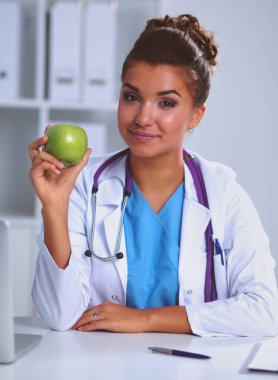 Kadın doktor, elinde yeşil bir elma, masada oturuyor.