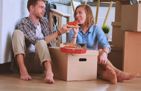 Pareja joven tiene una pausa para comer pizza en el suelo después de mudarse a una nueva casa con cajas a su alrededor. Pareja joven — Foto de Stock