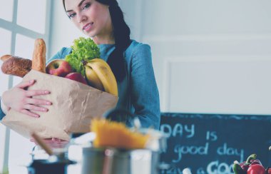 Sebzeli alışveriş poşeti taşıyan genç bir kadın. Mutfakta duruyor.