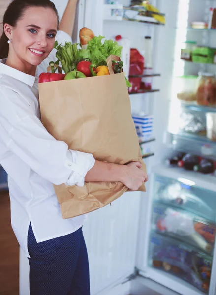 Jovem segurando bolsa de compras de supermercado com legumes. De pé na cozinha. Mulher na cozinha olhando para a câmera — Fotografia de Stock