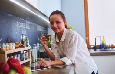 Genç bir kadın mutfakta tablet kullanıyor ve kahve içiyor..