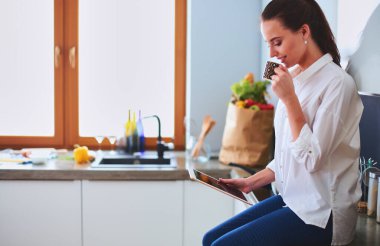 Genç bir kadın mutfakta tablet kullanıyor ve kahve içiyor..