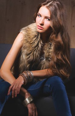 Mavi jeans giydiği siyah kanepede oturan zarif bir kadın portresi ve kürk yelek.