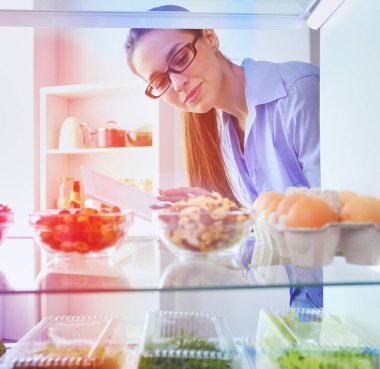 Sağlıklı yiyecek, sebze ve meyvelerle dolu açık buzdolabının yanında duran kadın portresi.