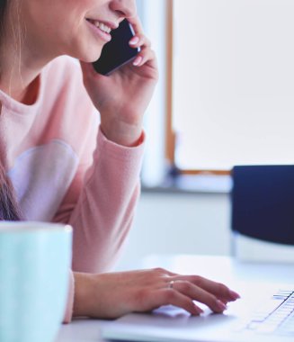 Genç kadın bir dizüstü bilgisayar kullanarak ve cep telefonuyla konuşurken mutfak masasında oturur