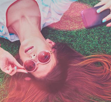 Güzel genç bir kadın yeşil çimenlerde yatarken telefonuyla selfie çekiyor. Güzel genç bir kadın selfie çekiyor..