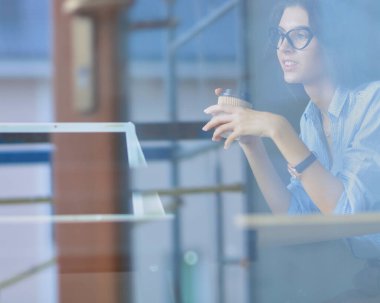 Ofis masasında dizüstü bilgisayarıyla oturan genç bir kadın. Pencereden bakıyor. Genç kadın..