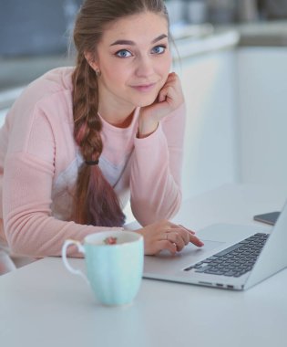 Genç kadın bir dizüstü bilgisayar kullanarak ve cep telefonuyla konuşurken mutfak masasında oturur