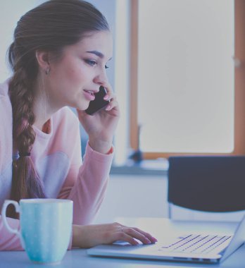 Genç kadın bir dizüstü bilgisayar kullanarak ve cep telefonuyla konuşurken mutfak masasında oturur