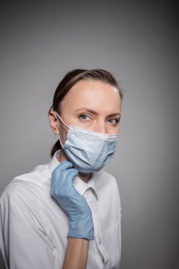 Yüzünde gri duvar arkaplanı olan tıbbi maskeli kadın doktor.