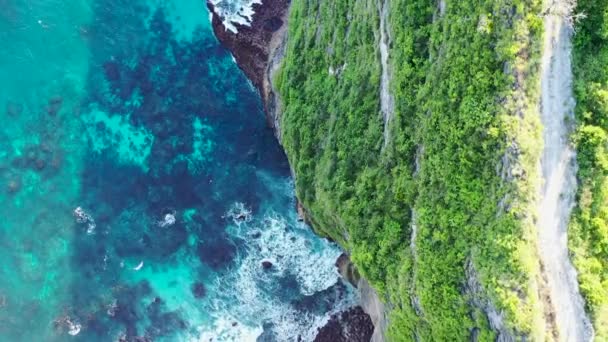 Filmische Luftaufnahmen von oben auf die vertikale Felswand. Drohne fliegt vorwärts mit Kamera nach unten und zeigt Fußweg, grüne Bäume und Meereswelle, die zu den Felsen spritzt — Stockvideo