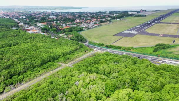 Letecký bezpilotní pohled na mangrovový les a rušnou silnici na mezinárodním letišti Bali. Letadla vzlétají. Na přistávací dráze se válí komerční letadlo. Letecké video mangrovníků na ostrově Bali, Indonésie — Stock video