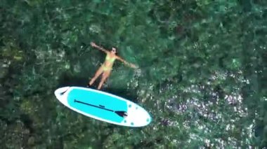 Gök mavisi deniz yüzeyinde uzanan güzel kadının havadan görünüşü. Sakin ve Rahat Kadın, Sup gemisinin yanında, sakin denizde dinleniyor. Komik yaz su sporları. Sağlıklı yaşam tarzı. Sörf, moda. Yaz tatili. 4K
