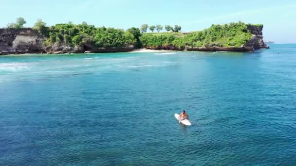 Un surfeur solitaire est assis sur sa planche de surf en attendant une vague comme un spec dans la vaste surface de l'océan. Vue aérienne de l'homme assis sur le surf et naviguant dans la magnifique baie de la mer tropicale pendant les vacances d'été — Video