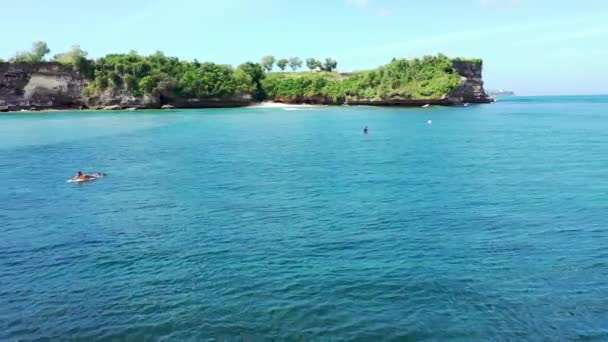 AERIAL: Surfistas flotando en el agua esperando las olas en línea por debajo de un alto acantilado rocoso. Resort turístico de lujo con bungalows, cabañas y hoteles rodeados de exuberante selva verde en Bali — Vídeos de Stock