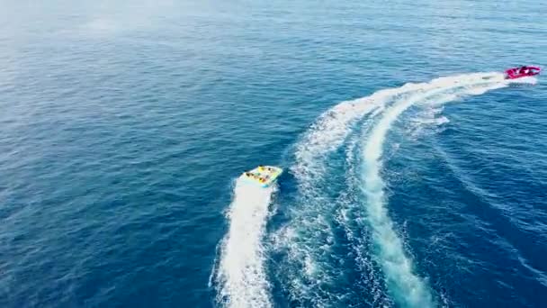 Σωληνωτή βάρκα με ανθρώπους να κινούνται πίσω από τη βάρκα στο νερό. Διασκέδαση στο νερό. Αεροφωτογραφία του κινητήρα ταχύτητα βάρκα τραβά κόκκινο φουσκωτά σχεδία καουτσούκ με τους ανθρώπους — Αρχείο Βίντεο