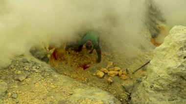 KAWAH IJEN, İNDONEZYA - 6 Haziran 2021. Sülfür madencisi, Ijen Yanardağı 'nın kraterinde zehirli sülfür gazı bulutlarıyla çevrili bir levyeyle çiğ sülfür bloklarını kırar.