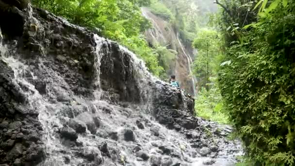 Um homem caminha pela cachoeira, olha em volta e levanta as mãos para cima, desfrutando de vista sobre a cachoeira na ilha de Java, Indonésia, Sem classificação de cores — Vídeo de Stock