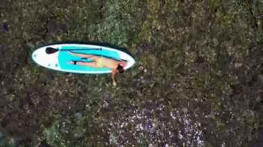 Genç bir kadının havadan çekilmiş görüntüsü bir kürek tahtasında uzanıyor ve berrak sudaki deniz mercanlarına bakıyor. Mavi denizde bir kürek tahtasının üzerinde uzanan seksi esmerin arka plan görüntüsü