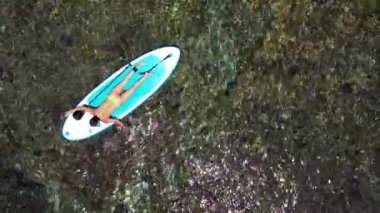 Genç kadın ayağa kalkmış SUP tahtasında yatarken dinleniyor. İnsansız hava aracıyla yapılan hava görüntüsü. Güzel deniz tabanı