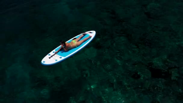 Αεροφωτογραφία μιας όμορφης γυναίκας ξαπλωμένης στη σανίδα του σερφ. Ηρεμία Χαλαρή Γυναίκα Ξεκουράζεται Στο Sup Surf. Ακραία καλοκαιρινά θαλάσσια σπορ. Υγιής τρόπος ζωής. Σέρφινγκ, Μόδα. Καλοκαιρινές διακοπές. 4K — Αρχείο Βίντεο