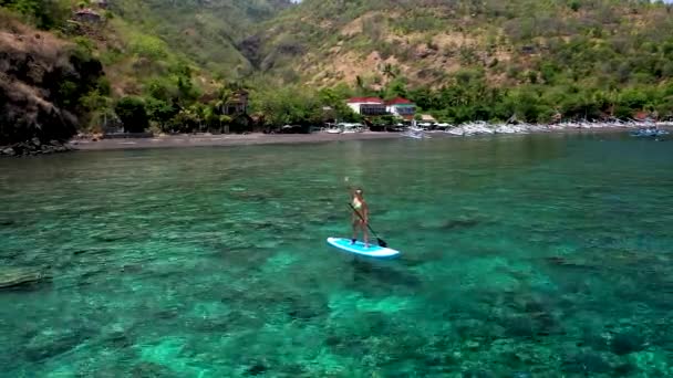 Młoda kobieta stojąca na desce i machająca ręką w powitaniu. Turkusowe morze z koralami w czystej wodzie. Strzał drona młodej kobiety z długimi włosami unoszącej się na wiosle — Wideo stockowe
