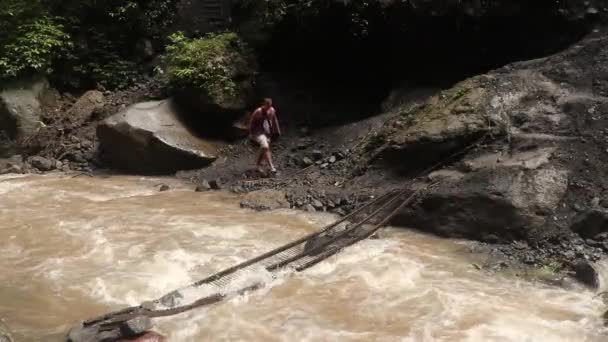 Turista átkel egy vad folyón egy instabil gyaloghídon. Régi acélhíd a hegyi folyón át — Stock videók