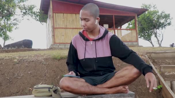 Ένας νεαρός Ινδονήσιος ακούει μουσική με ακουστικά. Ασιάτης κάθεται, καπνίζει και παίζει smartphones στο πάρκο — Αρχείο Βίντεο
