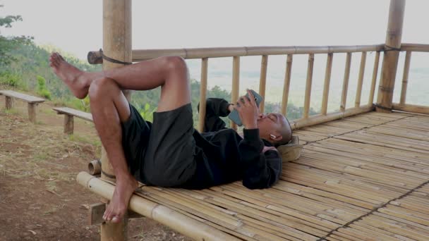 アジア人の男がガゼボに寝そべってた。竹のガゼボで休んでいる男 — ストック動画